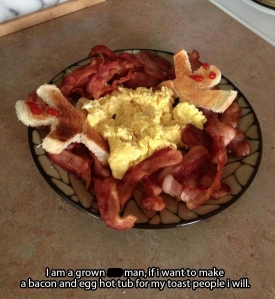 A bowl with lots of bacon around the outside, scrambled eggs in the middle, and toast cut into the shapes of men with ketchup smiley faces. "Bacon and egg hot tub for toast men" 
